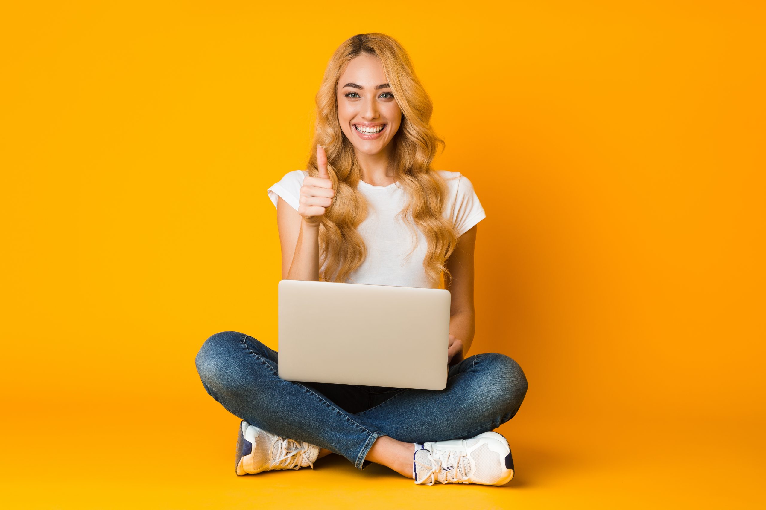 Cheerful woman using laptop and showing thumb up on yellow studio background, smiling to camera